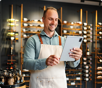 Man wearing an apron standing in front of a wine rack while looking at a tablet