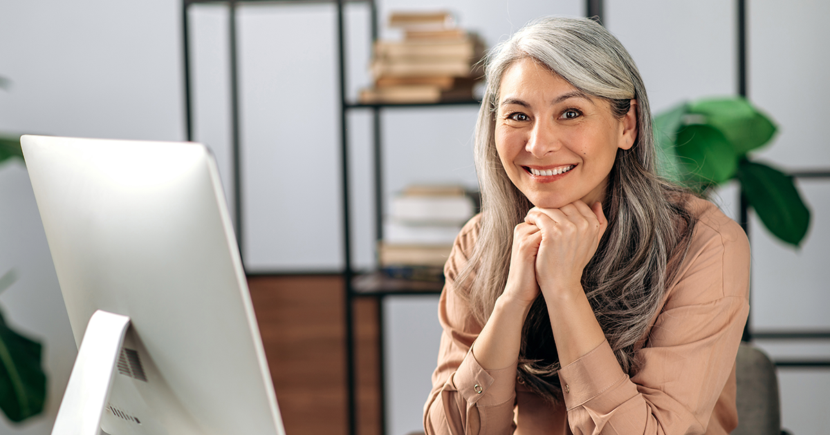 Business woman sitting at desk with computer resting chin on hands looking at camera while smiling