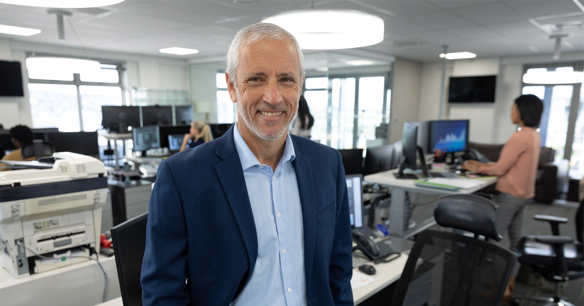   Save Download Preview Portrait of senior Caucasian businessman working in a modern office, standing and smiling to camera, colleagues in the background.