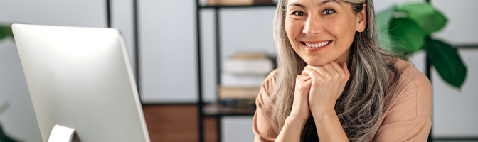 Business woman sitting at desk with computer resting chin on hands looking at camera while smiling