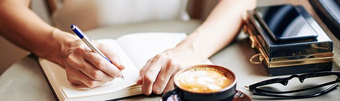 Closeup of a person writing in a journal with a cup of coffee and glasses on a table.