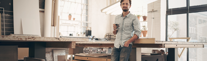 young male designer standing comfortably in his naturally lit workshop, holding his morning cup of coffee and smiling at the camera