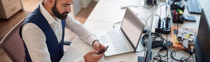 Young business man working in office texting on cell phone while working on computer.