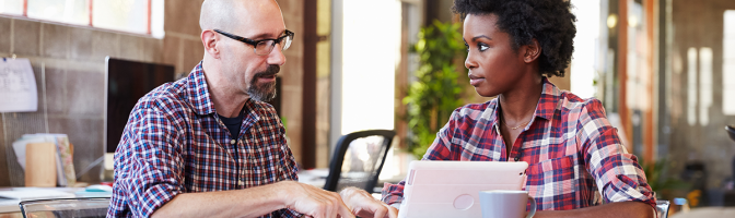 Two professionals sit at meeting table working on digital tablet
