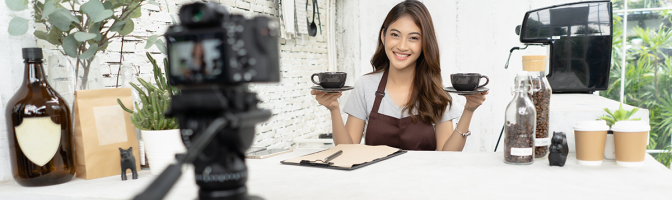 young female barista in apron smiling with recording video camera filming at coffee shop counter.