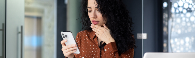 Shocked  young business woman sitting in office at desk with laptop, holding phone, hand on face