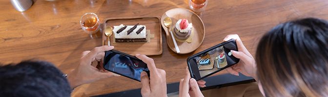 Two people take a photo with their mobile phones of a plate of dessert and beverage.