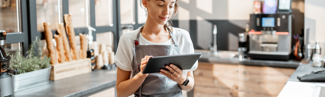Young saleswoman working with a digital tablet at the counter of cafe or confectionary shop.