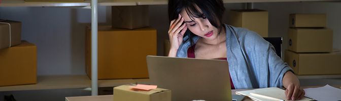 Woman holding her head while looking at a laptop with boxes on shelves behind her.