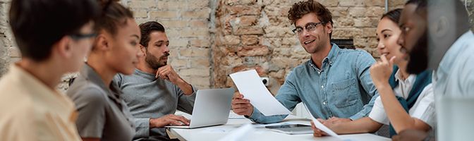 A group of six business professionals have a discussion around a meeting table.