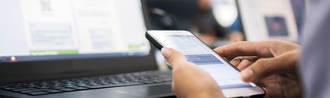 Close up of a person typing into a smart phone in front of a laptop.