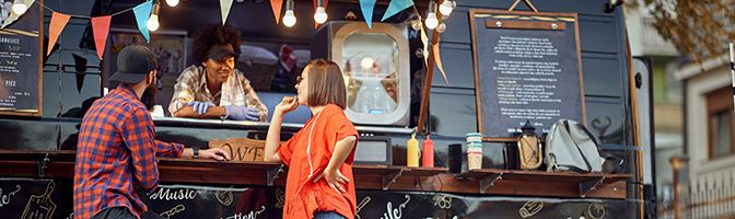 A chef entrepreneur has a friendly conversation with two customers while serving them through the window of her food truck.