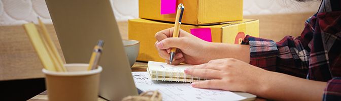 Close up of a small business owner writing in a notebook at a desk in front of a laptop.