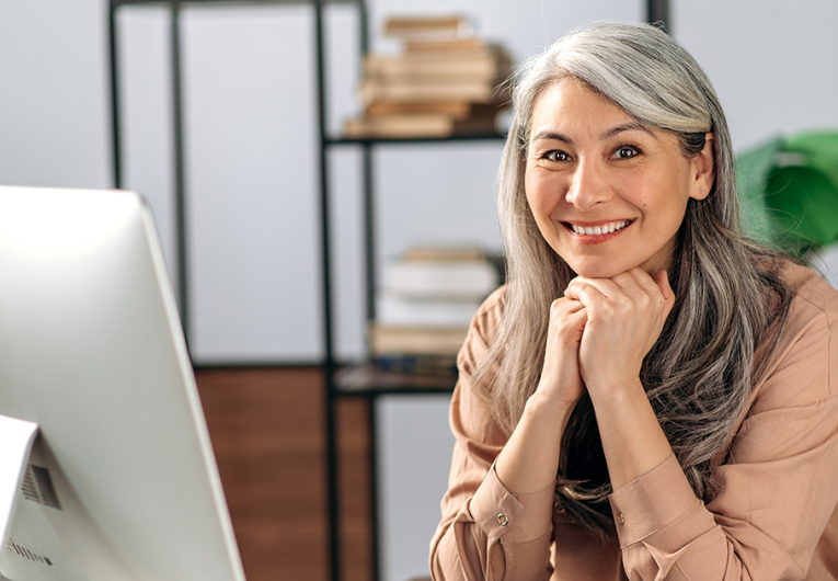 Business woman sitting at desk with computer resting chin on hands looking at camera while smiling