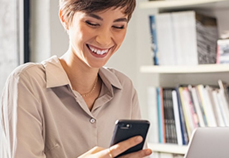 Entrepreneur smiling while looking at mobile device in front of a laptop as she sits at a desk.