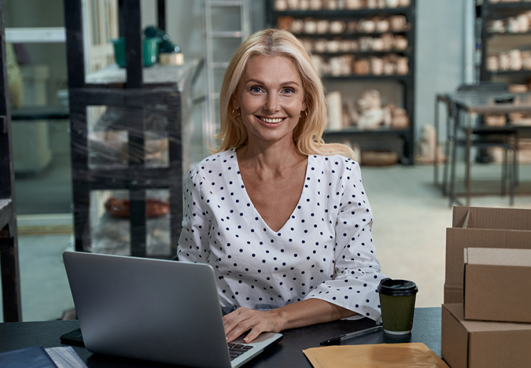 Happy female business owner or business woman sitting in her art studio or craft pottery shop with handmade ceramic products, working on laptop and looking at camera