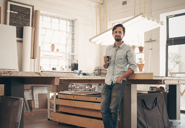young male designer standing comfortably in his naturally lit workshop, holding his morning cup of coffee and smiling at the camera