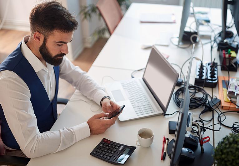 Young business man working in office texting on cell phone while working on computer. 