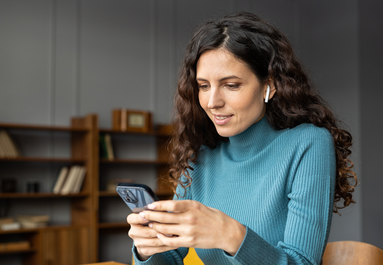 Smiling woman sitting at office desk looking at phone with earbuds in ears listening to podcast. 