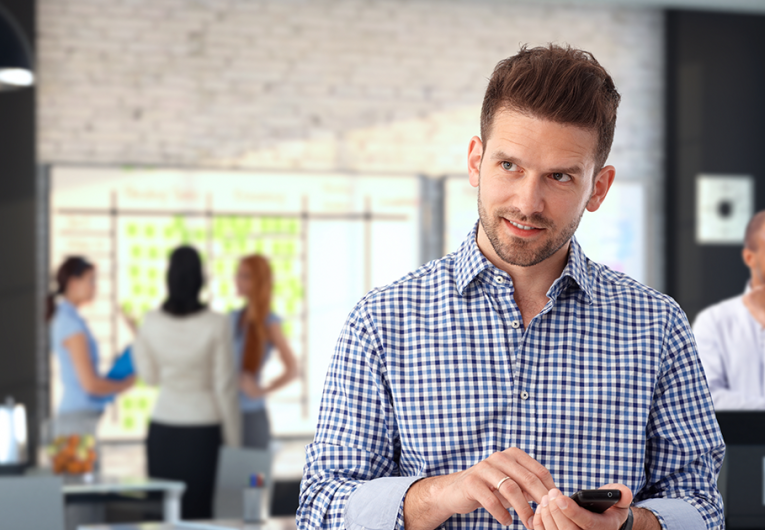 Casual businessman using mobile phone at modern stylish office, smiling. Employees working and talking in the background. 