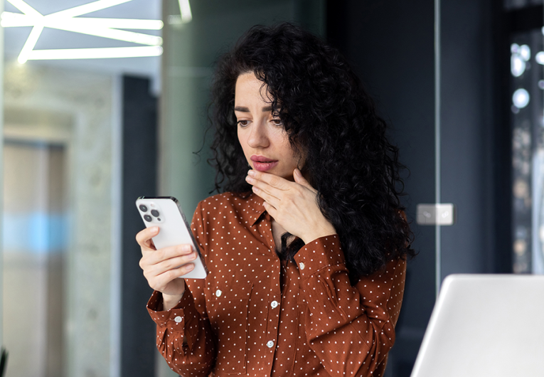 Shocked  young business woman sitting in office at desk with laptop, holding phone, hand on face