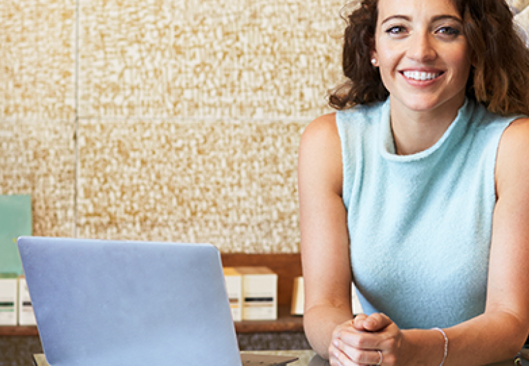 Young woman working in clothes shop leaning on counter with open laptop. 