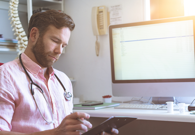 Side view of a Caucasian male doctor checking medical paper in clinic against his computer in background