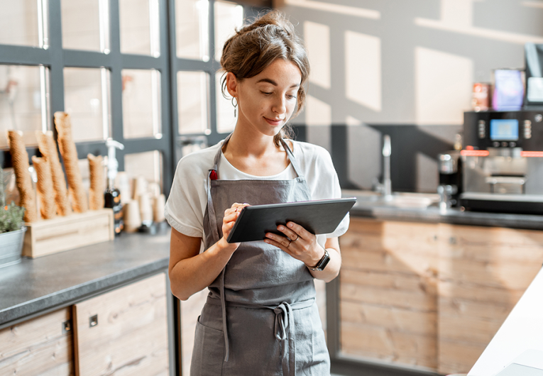 Young saleswoman working with a digital tablet at the counter of cafe or confectionary shop.