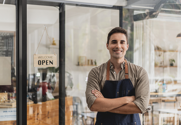 A male cafe employee standing in front of the store greets customers and has a sign that says open to show that the shop is open,