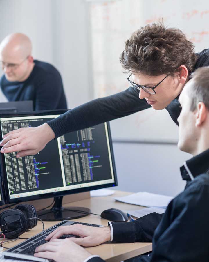 A man gestures at a computer screen while another sits at the keyboard