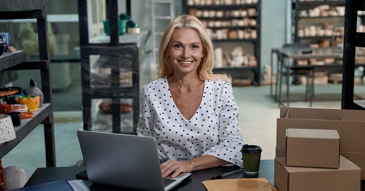 Happy female business owner or business woman sitting in her art studio or craft pottery shop with handmade ceramic products, working on laptop and looking at camera