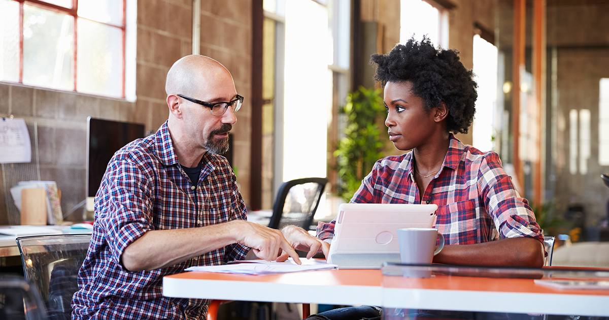 Two professionals sit at meeting table working on digital tablet