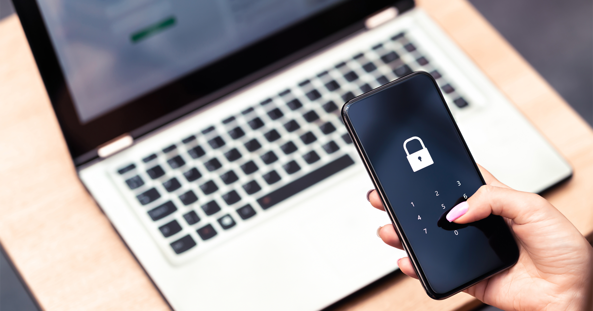 Hand holding a phone hovering over laptop computer with a password lock on the screen. 