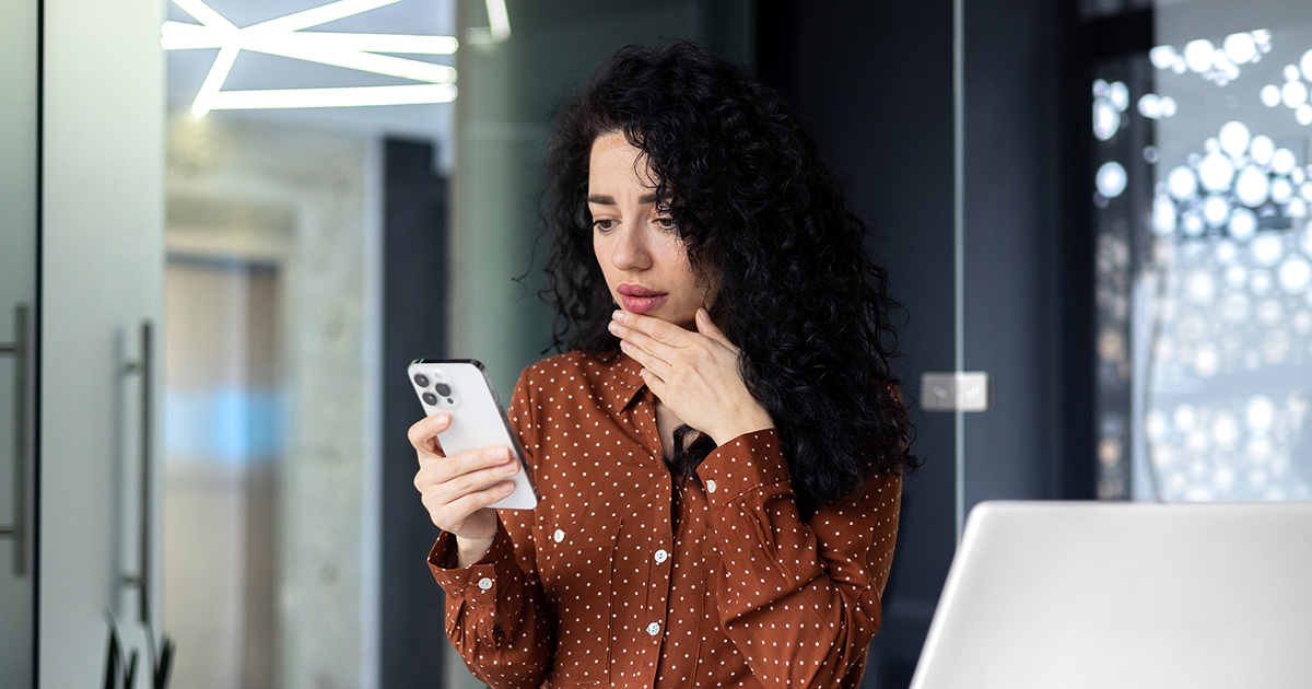 Shocked  young business woman sitting in office at desk with laptop, holding phone, hand on face