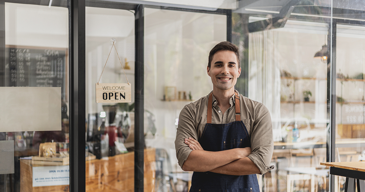 A male cafe employee standing in front of the store greets customers and has a sign that says open to show that the shop is open,