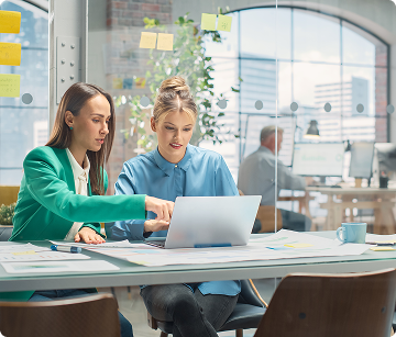 Two women sitting at a table looking at a laptop in a large office area